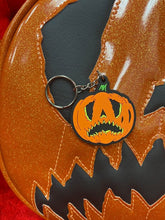 Load image into Gallery viewer, Crying pumpkin keychain in orange and black  on top of the bag in the same design. 