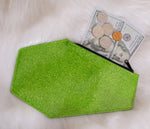 Green glitter coffin shaped coin bag with green stitching and a black zipper with two Hundred dollar bills, three quarters a nickel and a penny sticking out of it 