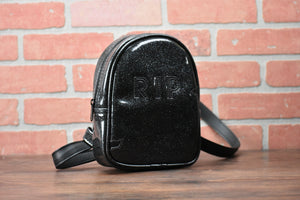 Black glitter small headstone back pack with "RIP" across the front.  Photographed in front of brick wall. 