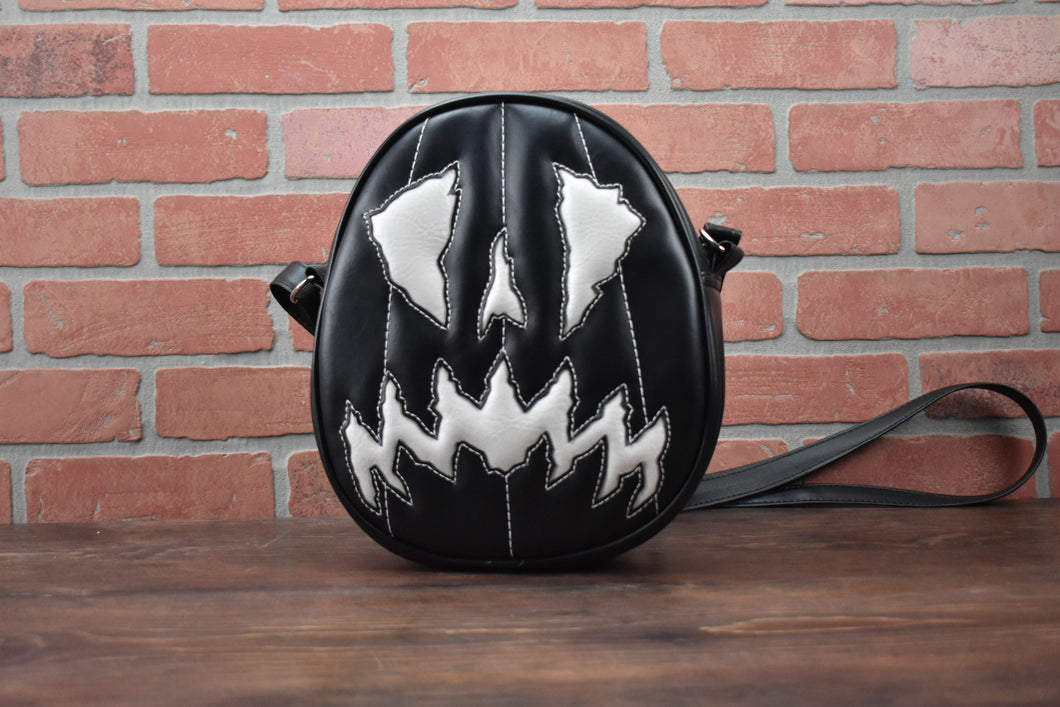 Black and white scaredy-cat face oval pumpkin , white stitching detail on the face handbag. Against a brick wall. 