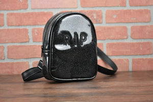 Black glitter small headstone back pack with "RIP" across the front.  Photographed in front of brick wall. 